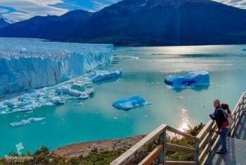 Fotografia do turismo no Glaciar Perito Moreno, pessoas em uma estrutura de metal que dá vista para o Lago Argentino, ao fundo da imagem um gigantesco glaciar emerge das águas.
