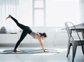 Uma mulher em uma sala fazendo uma posição de yoga: palmas das mãos e um pé apoiado do chão e um dos pés pra cima.