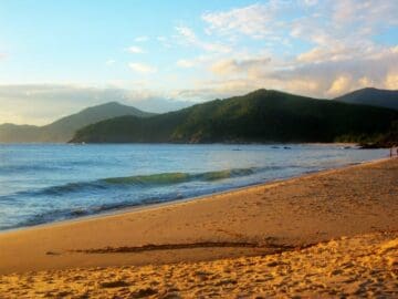 Praia arenosa com faixa extensa de areia clara, ondas suaves quebrando na orla e horizonte azul ao fundo.