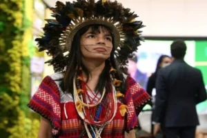 Txai Suruí linda usando um grande cocar e roupas indígenas em evento na França