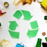 Imagem representando resíduos e reciclagem: símbolo verde de reciclagem ao centro, cercado por diversos tipos de resíduos como vidro, plástico, metal, pilhas e restos orgânicos.