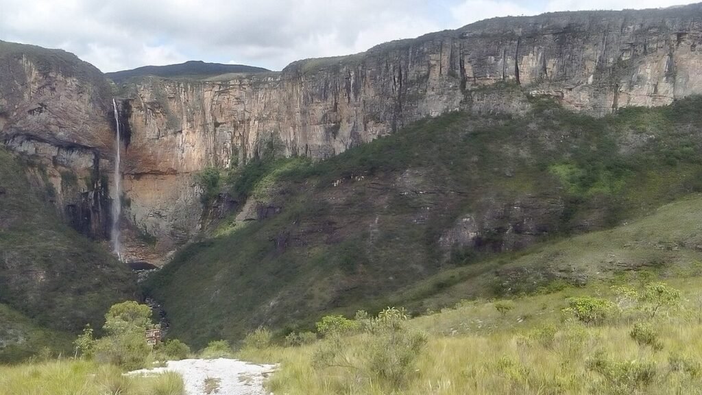Grande paredão de pedra com uma cachoeira alta e fina caindo no vale, com campo em primeiro plano