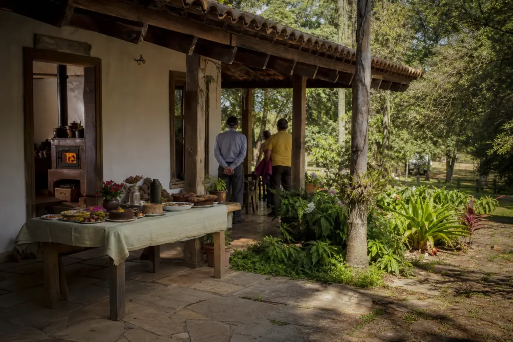 Varanda de casa de fazenda antiga com fogão a lenha e mesa de quitutes, visitantes de costas: turismo cultural rural