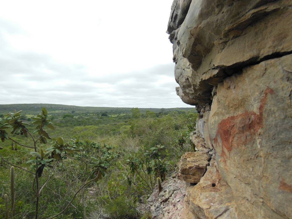 Paredão de arenito com pintura rupestre avermelhada, ao lado de uma vista da caatinga